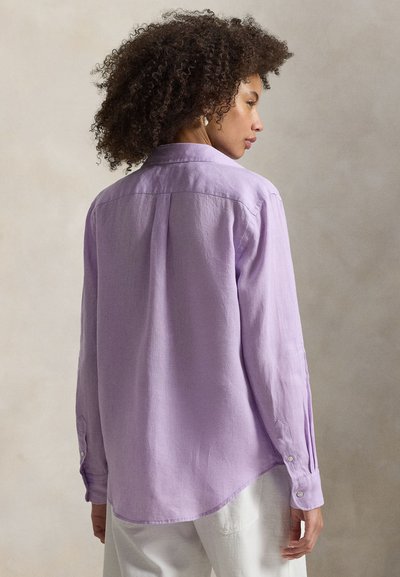 Γυναίκα με σγουρά μαλλιά που φοράει πουκάμισο με μακριά μανίκια σε λεβαντίνια απόχρωση και λευκό παντελόνι, στραμμένη στο προφίλ, μπροστά σε ουδέτερο μονόχρωμο φόντο.