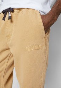 Beżowe bawełniane spodnie z elastycznym pasem i brązowym sznurkiem do regulacji. Posiadają haftowane logo "TOMMY JEANS" w tym samym kolorze po bokach.