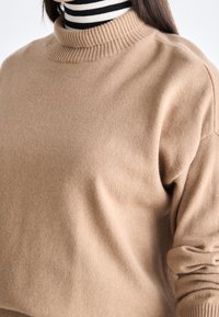 Pull en laine brun clair avec un col montant côtelé et des manches longues, présentant une texture douce et lisse ainsi qu'un motif subtil au niveau des poignets.