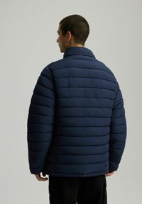 Nadmuchiwany granatowy kurtka z kołnierzykiem stojącym, z poziomym pikowaniem dla tekstury i luźnym krojem. Brak widocznych zapięć.