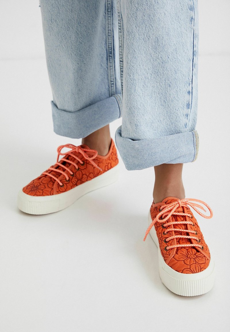 Personne portant un jean bleu clair retroussé et des baskets orange à motifs floraux avec des semelles blanches et des lacets orange.