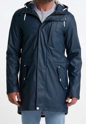 Homme portant un imperméable bleu marine avec poches à rabat boutonnées et capuche sur une chemise grise et un jean bleu clair.