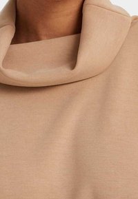 Maglia a collo alto in tessuto beige morbido, con collo ripiegato e superficie liscia e testurizzata. Design semplice e minimalista.