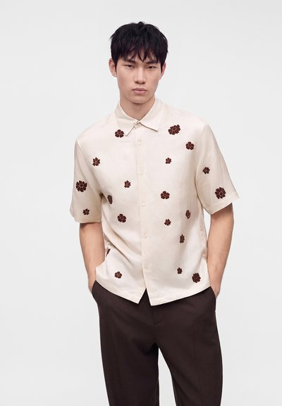 Jeune homme portant une chemise beige à manches courtes avec des imprimés floraux marron foncé et un pantalon marron foncé, debout les mains dans les poches.