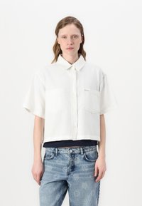 White cropped shirt with a collar, short sleeves, and two chest pockets, made of a lightweight fabric, paired with faded blue jeans.