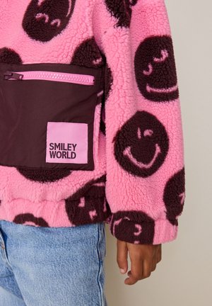 Veste polaire rose avec des motifs de visages souriants marron, poche avant zippée marron foncé et étiquette "SMILEY WORLD", portée avec un jean bleu clair.