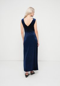 Γυναίκα με μακριά ξανθά μαλλιά, φορώντας ένα αμάνικο νυφικό βαθύ V-πλάτης ναυτικό μπλε βελούδινο maxi φόρεμα και μαύρες γάτες τακούνια, γυρισμένη προς τα πίσω.