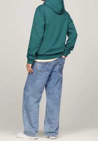 Sudadera verde bosque con capucha ajustable, combinada con unos vaqueros de mezclilla anchos de color azul claro. Zapatillas blancas completan el look.