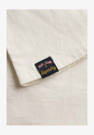 Rogówka białej bawełnianej koszuli z granatową metką, na której znajdują się dwie flagi oraz wypisane złotą nitką "Superdry". Gładka tekstura z zagiętym brzegiem.