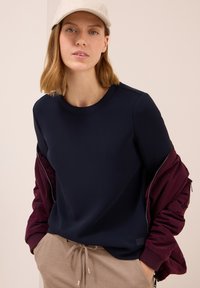Tummansininen sweatshirt, jossa on pyöreä kaula-aukko, yhdistettynä viininpunaisiin bomber-takkeihin. Tausta on vaalean beige. Rentoa tyyliä elastisella vyötäröllä.