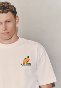 Maglietta bianca in cotone con grafica arancione colorata e foglie verdi. Il testo recita "EL SOL NARANJA" con dettagli sulla località in nero.