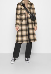 Rutigt ullrock i beige och svart, dubbelknappad design med fickor. Kombinerat med mörka jeans och svarta sneakers, bärande en liten svart väska.