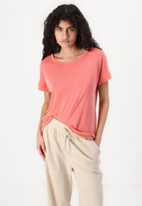 T-shirt corallo con maniche corte e scollo rotondo, abbinata a pantaloni beige chiaro con coulisse, su uno sfondo bianco uniforme.