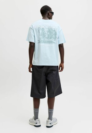 Muž v svetlomodrom tričku s grafikou, čiernych krátkych nohaviciach po kolená, sivých ponožkách, bielych teniskách a slnečných okuliaroch, stojaci chrbtom k bielemu pozadiu.