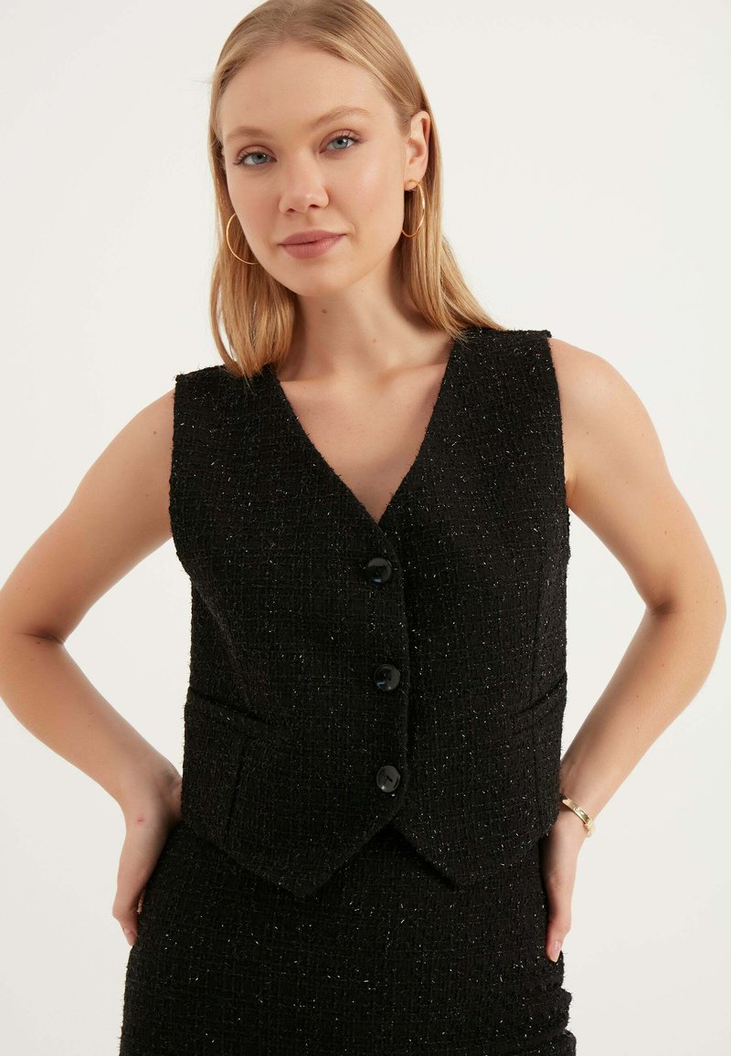 Svart ärmlös väst gjord av textilerat tyg med en subtil glans, med v-ringning, två framfickor och tre svarta knappar.