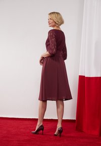 Robe bordeaux avec des manches en dentelle, jupe évasée et taille cintrée. Assortie à des chaussures à talons brillantes sur un fond de tapis rouge.