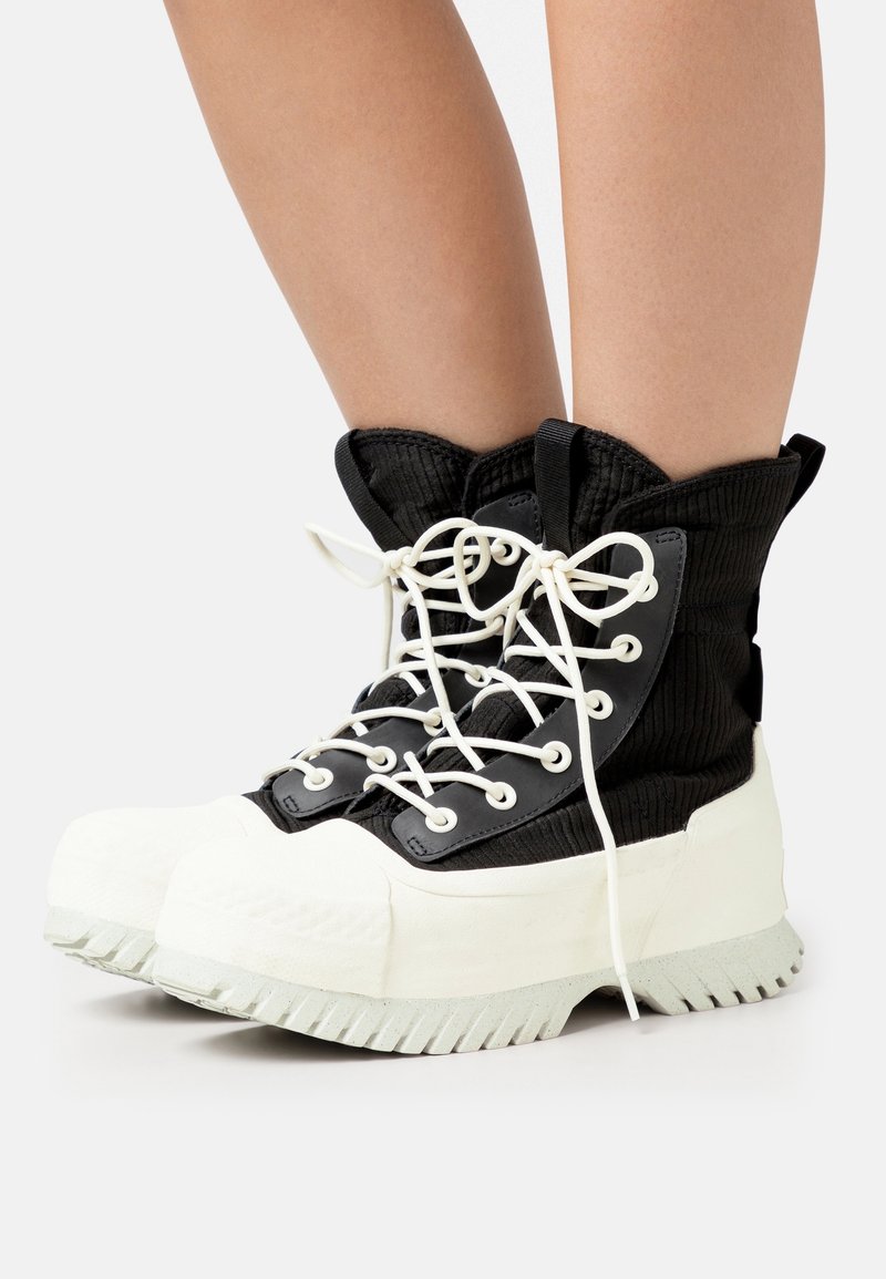 Jambes portant des bottes montantes en velours côtelé noir avec des semelles épaisses en caoutchouc blanc et des lacets blancs noués en nœud, sur un fond blanc.