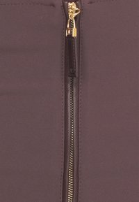 Dettaglio di tessuto borgogna con una cerniera tono oro dotata di un tirante in pelle, posizionata centralmente. Finitura liscia e testurizzata attorno alla cerniera.