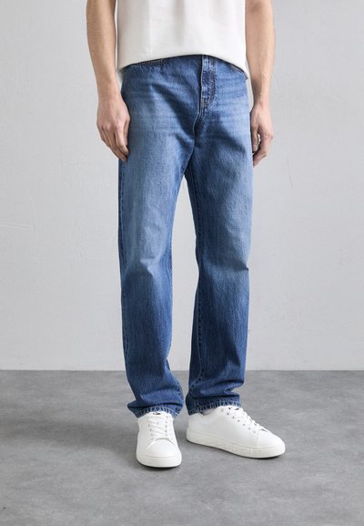 Jean en denim bleu clair avec une coupe droite, présentant des coutures visibles et un design à cinq poches, associé à des baskets blanches.
