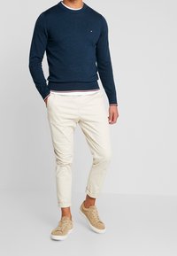 Jersey de punto azul marino con cuello redondo, combinado con pantalones chinos color crema y zapatillas de ante beige. Presenta un logo sutil en el pecho.