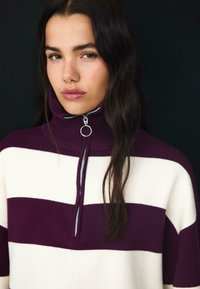 Pull à col roulé rayé avec de larges rayures horizontales en violet profond et blanc, avec une fermeture à demi-glissière et une texture de col côtelé.