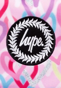 Pyöreä musta brodeerattu merkki, jossa on "hype."-teksti, ympäröity valkoisilla laakerinlehdillä, värikkään vaaleanpunaisen taustan päällä, jossa on sydänkuvioita.