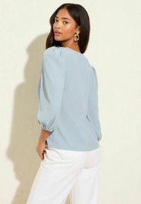Femme aux longs cheveux noirs portant un haut bleu clair à manches bouffantes et un pantalon blanc, posant contre un mur clair uni.