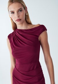 Vīna sarkanas kleitas ar drapētu kakla izgriezumu, asimetrisku plecu un savilkumu detaļām. Gluds audums, piegulošs siluets, kas izceļ vidukli.