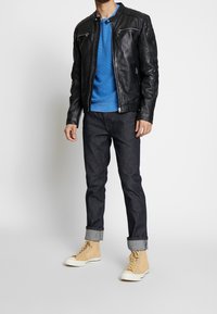 Zwarte leren jack met ritszakken, blauwe gebreide trui, donkere spijkerbroek en beige high-top sneakers met witte zolen.