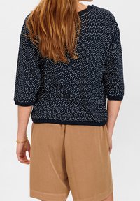 Marineblaue Sweatshirt mit weißen Punkten, dreiviertellangen Ärmeln, rundem Ausschnitt und geripptem Saum, kombiniert mit beigefarbenen Shorts.