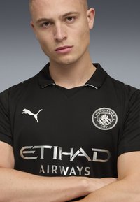 Svart fotbollströja med V-ringning, med en vit Puma-logotyp, silvertexten "ETIHAD AIRWAYS" och Manchester Citys emblem på vänster sida.
