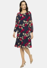 Granatowa sukienka w kwiaty z przezroczystymi, długimi rękawami, z czerwonymi i beżowymi kwiatami. Kształt A-linii, do kolan, wykonana z lekkiego materiału. Elegancki okrągły dekolt.