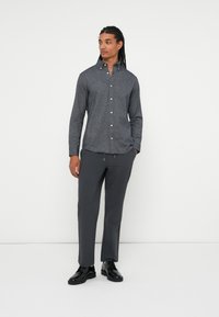 Bărbat cu păr lung împletit, purtând o cămașă cu butoni gri închis, pantaloni gri închis și pantofi eleganți negri, stând cu mâinile în buzunare.