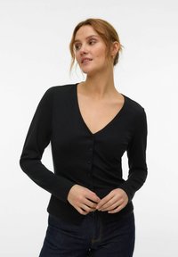 Femme aux cheveux châtain clair portant un haut noir à manches longues avec un col en V boutonné et un jean foncé, ajustant ses mains au niveau de la taille.
