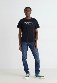 Sort bomulds t-shirt med hvid "Pepe Jeans London" logo, parret med blå denim jeans og grønne og hvide sneakers. Enkelt fit, afslappet stil.