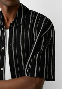 Svart och vit randig kortärmad skjorta gjord av textil med struktur, med krage och knappstängning fram. Vit undertröja synlig.