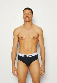 Smiling young man standing against a plain background, wearing black briefs with a white waistband and "ellesse" logo.