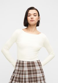 Γυναίκα με κοντά σκούρα μαλλιά, φορώντας λευκό μπλουζάκι με μακριά μανίκια και καρό καφέ φούστα, ποζάροντας με τα χέρια στη μέση μπροστά σε λευκό φόντο.