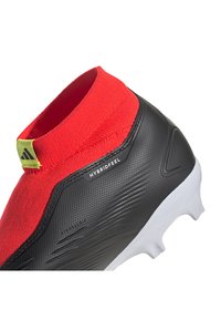 Botas de fútbol con un cuello de punto rojo y un cuerpo negro texturizado. Incluyen una suela blanca y detalles de marca en el lado.