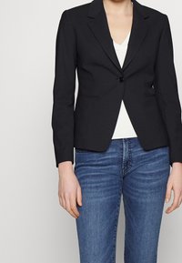 Černý přizpůsobený blazer s jedním knoflíkem, klopami a dvěma předními kapsami. Nosený přes bílý top, v kombinaci s modrými džíny.