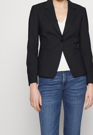 Blazer noir ajusté avec une fermeture à un bouton, revers crantés et deux poches avant. Porté par-dessus un haut blanc, associé à un jean bleu.
