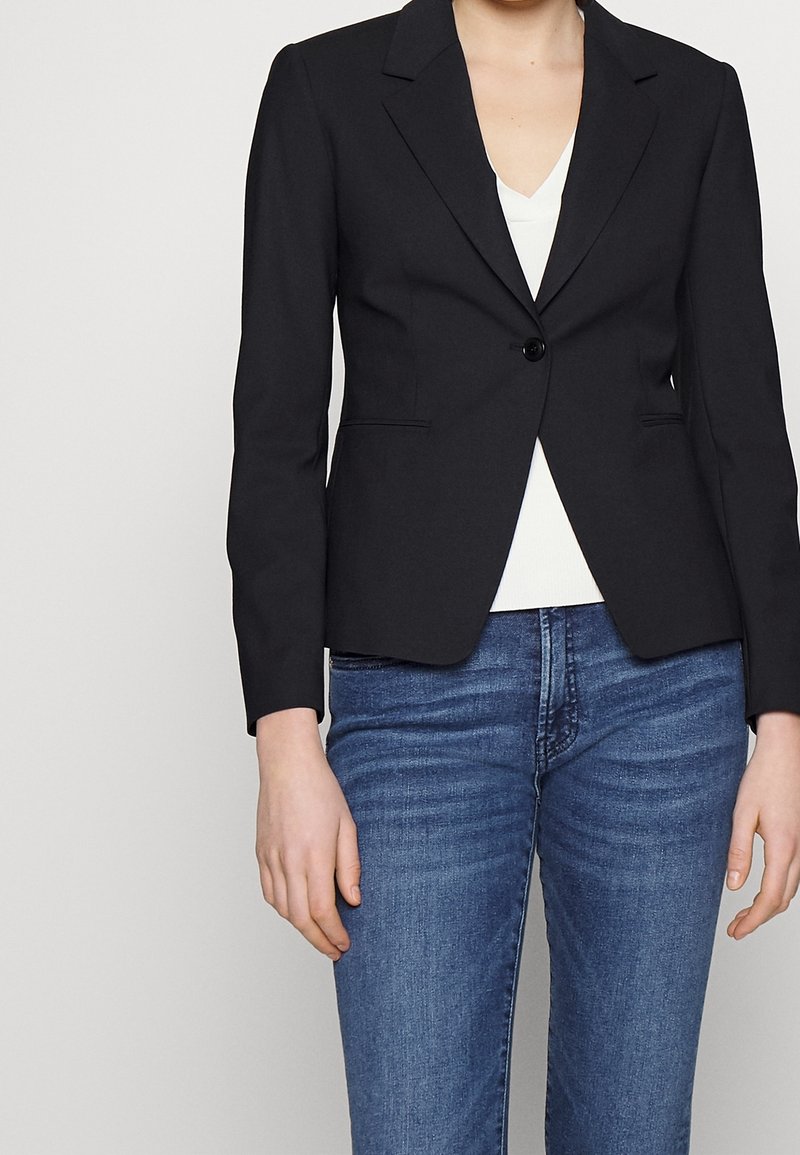 Černý přizpůsobený blazer s jedním knoflíkem, klopami a dvěma předními kapsami. Nosený přes bílý top, v kombinaci s modrými džíny.
