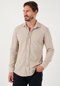 Camisa de botones de manga larga beige con una textura suave, cuello y dobladillo redondeado. Presenta botones marrón claro y un ajuste relajado.