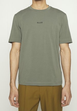 Mann trägt ein olivgrünes BOSS-T-Shirt mit kurzen Ärmeln und senffarbene Hose, steht vor einem schlichten, hellen Hintergrund.