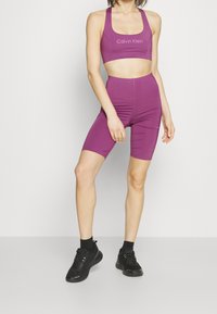 Conjunto de top deportivo corto morado y pantalones cortos de ciclismo, hecho de tela elástica, con el logo de Calvin Klein en la parte superior. Combinado con zapatillas negras.