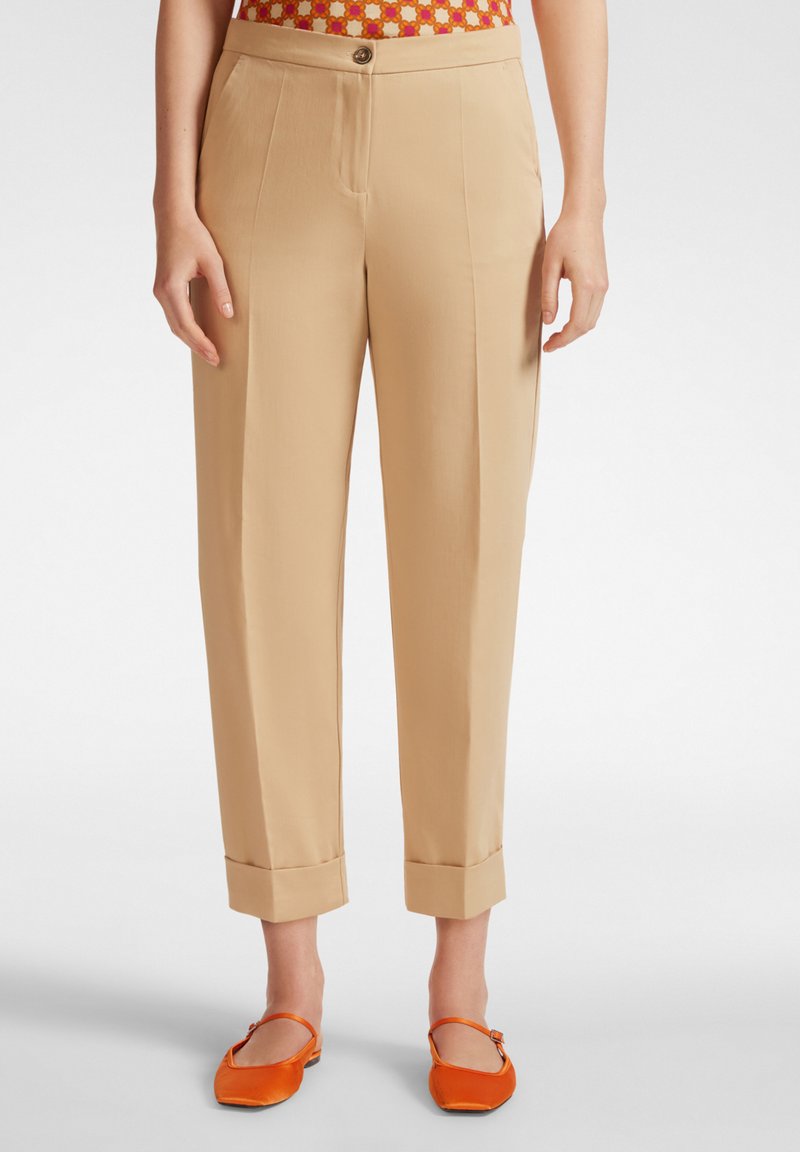 Pantaloni beige su misura con taglio dritto, orli arricciati e chiusura a bottone in vita. Indossati con scarpe flat arancioni.