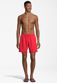 Röda badbyxor med resårmidja och snörning, med en logotyp på nedersta högra hörnet. Bärs med svarta flip-flops.