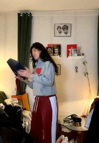 Jong persoon met krullend haar die een boek leest in een gezellige kamer met planken, ingelijste foto’s en een hoge plant naast een glazen tafel.