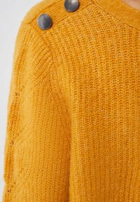 Manche de pull tricoté jaune moutarde avec deux boutons métalliques ronds près de la couture de l'épaule.