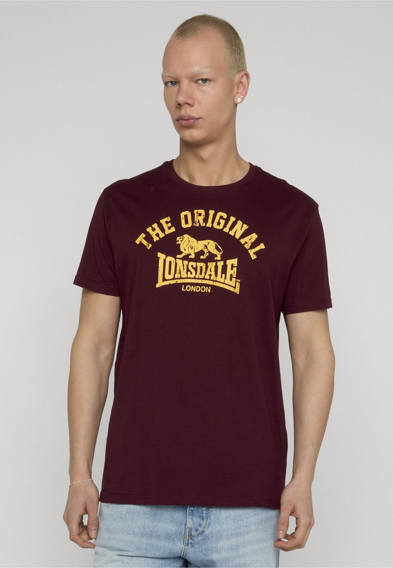 Jaunas vyras su trumpais blondinų plaukais, vilkintis bordo spalvos "Lonsdale London" marškinėlius ir šviesiai mėlynas džinsus, stovintis priešais paprastą foną.